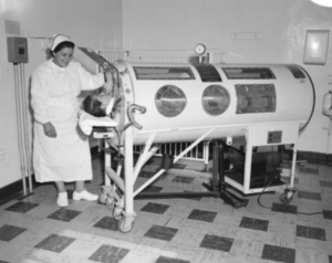 Black and white photo shows a nurse attending to a boy in an iron lung.