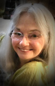 Portrait of Martha Lillard in 2016,. Smiling, wearing glasses, blond hair and a yellow shirt.